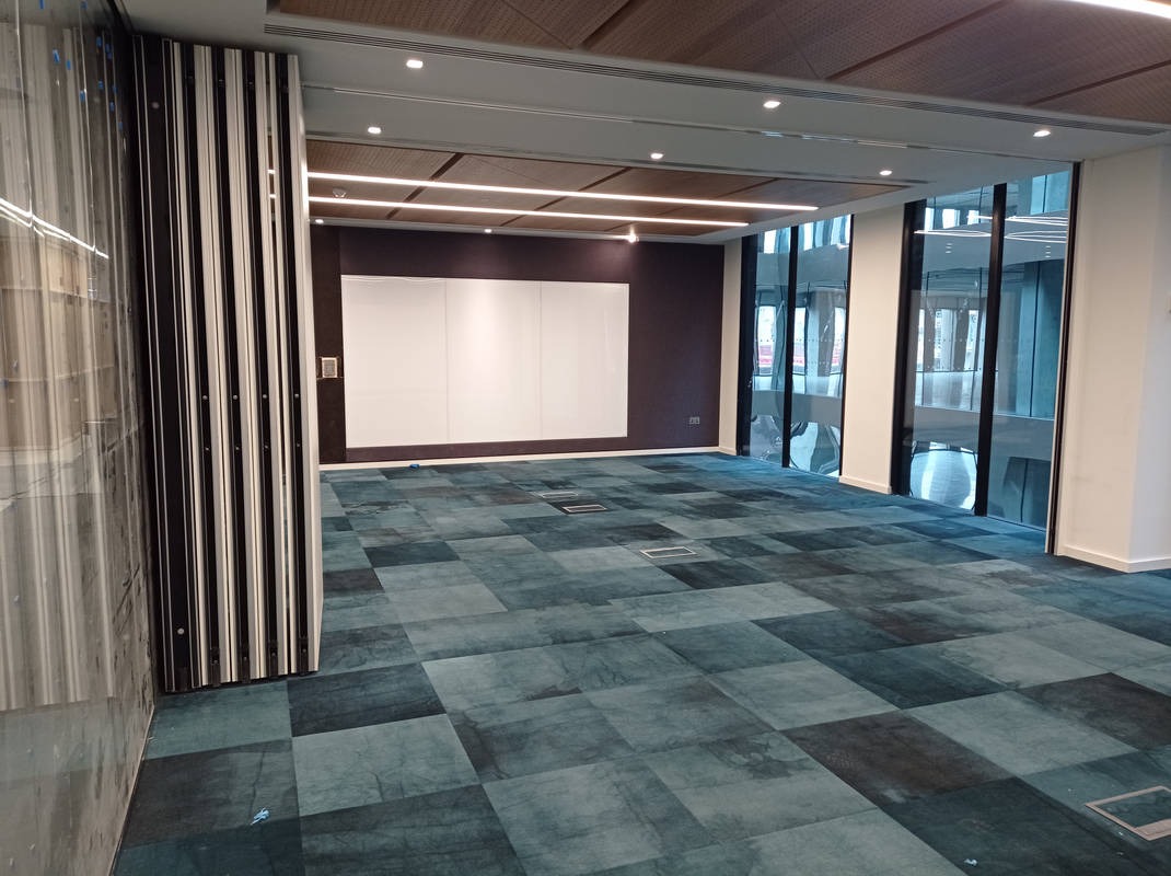 Wykończone wnętrze biurowe z wykładziną dywanową w odcieniach błękitu i szarości, widok na białą tablicę i okna. Nowoczesne oświetlenie sufitowe.