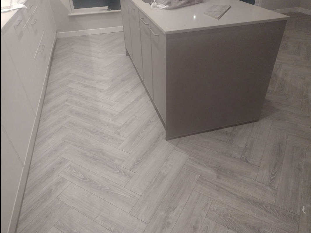 Jasnoszara podłoga ułożona w jodełkę w kuchni, widoczna wyspa kuchenna i białe szafki. Minimalistyczny design, subtelne oświetlenie.