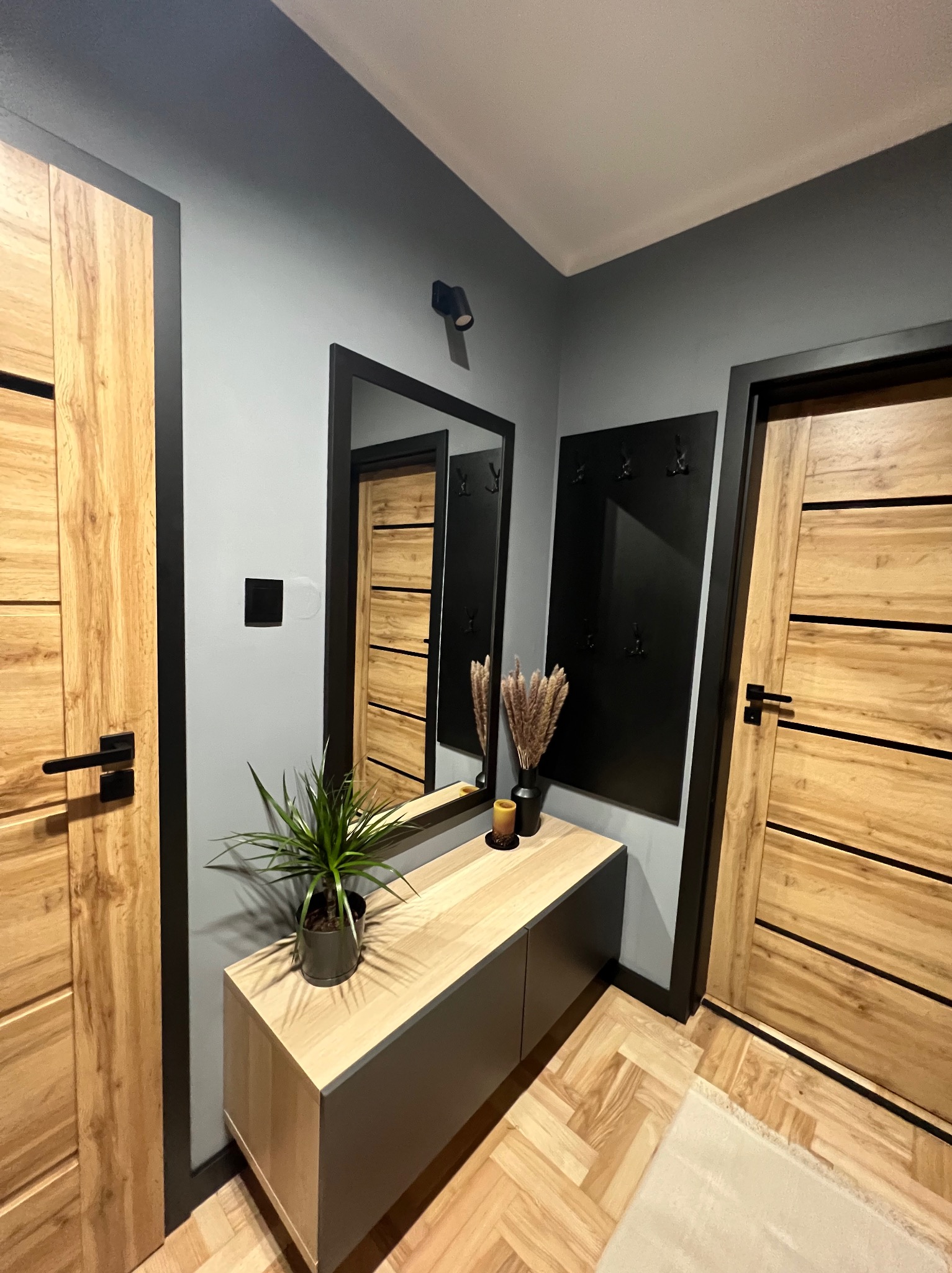 Stylowy przedpokój z drewnianymi drzwiami, lustrem w czarnej ramie, szafką i wieszakiem. Na podłodze parkiet w jodełkę, szare ściany i roślina doniczkowa.