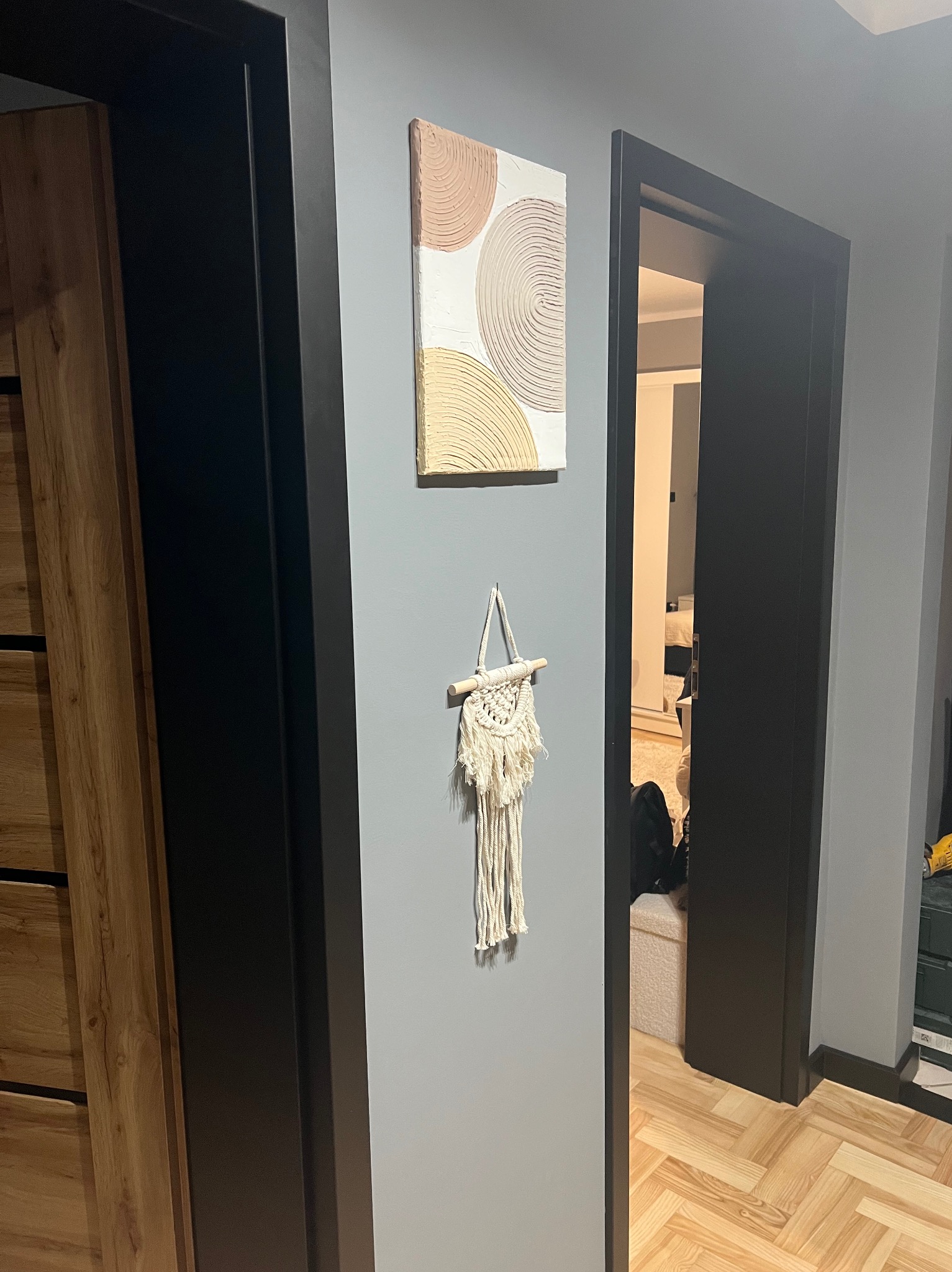 Nowoczesne wnętrze: ciemna rama drzwi, drewniana podłoga w jodełkę, abstrakcyjny obraz i makrama na szarej ścianie. Widok na pokój przez otwarte drzwi.