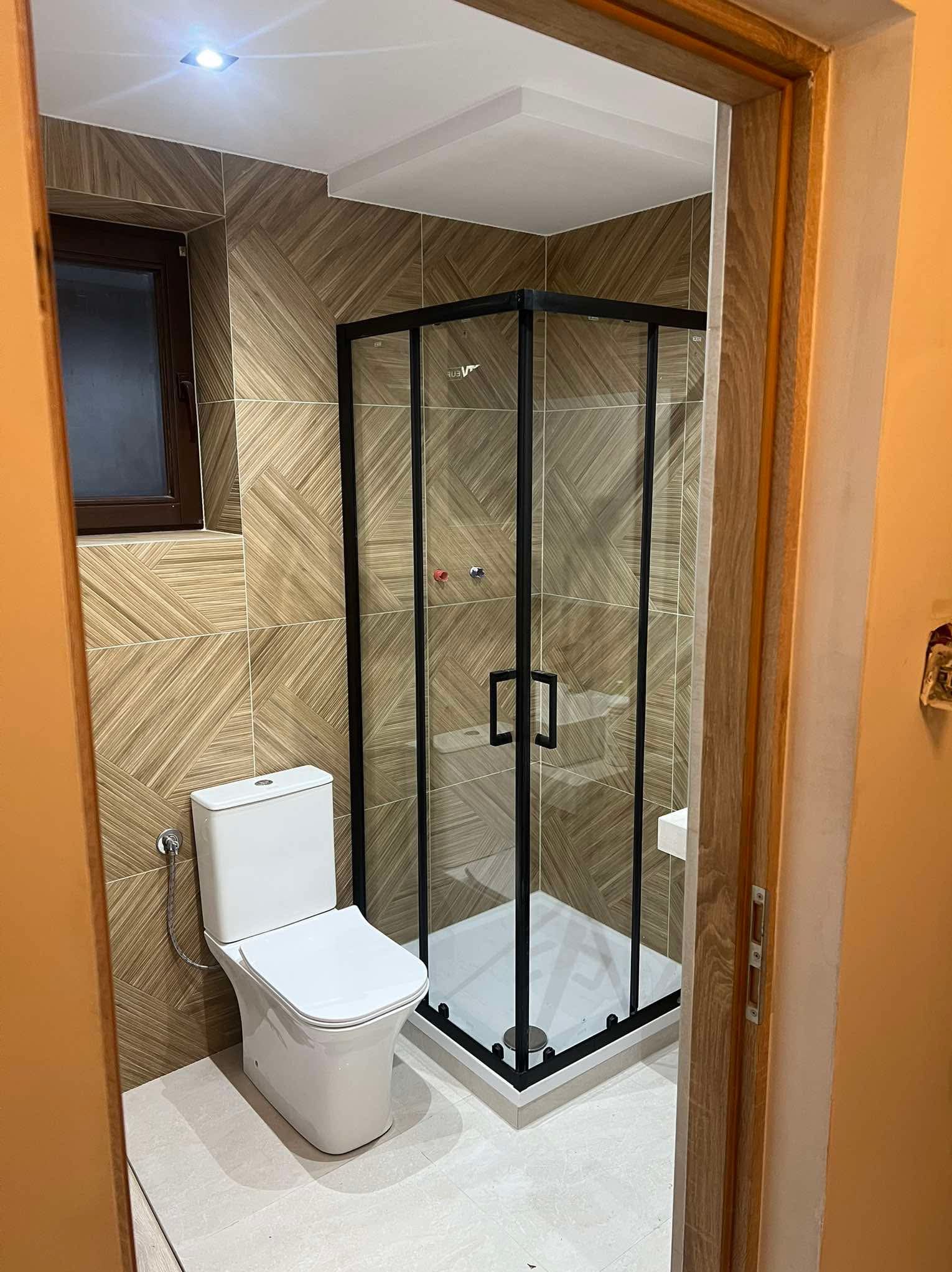 Nowoczesna łazienka po remoncie: kabina prysznicowa z czarną ramą, wisząca toaleta i płytki imitujące drewno. Widok zza futryny drzwi.