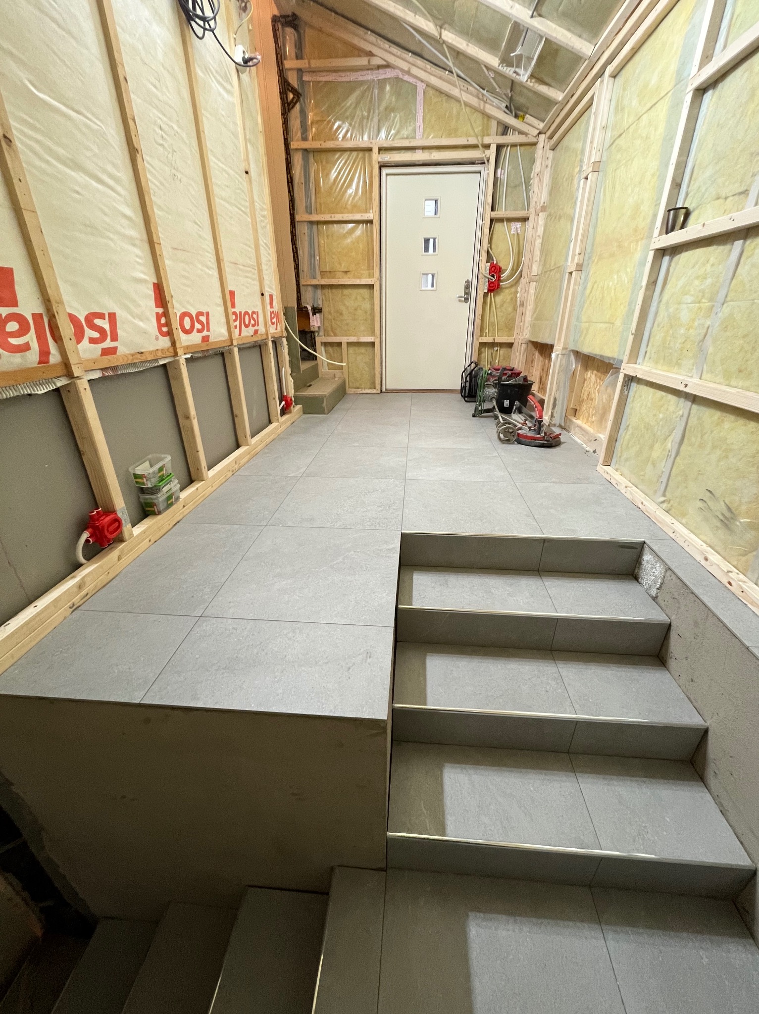 Wykończone płytki podłogowe i schody w surowym wnętrzu z izolacją. Widoczne narzędzia i materiały budowlane. Etap prac wykończeniowych w nowym domu.