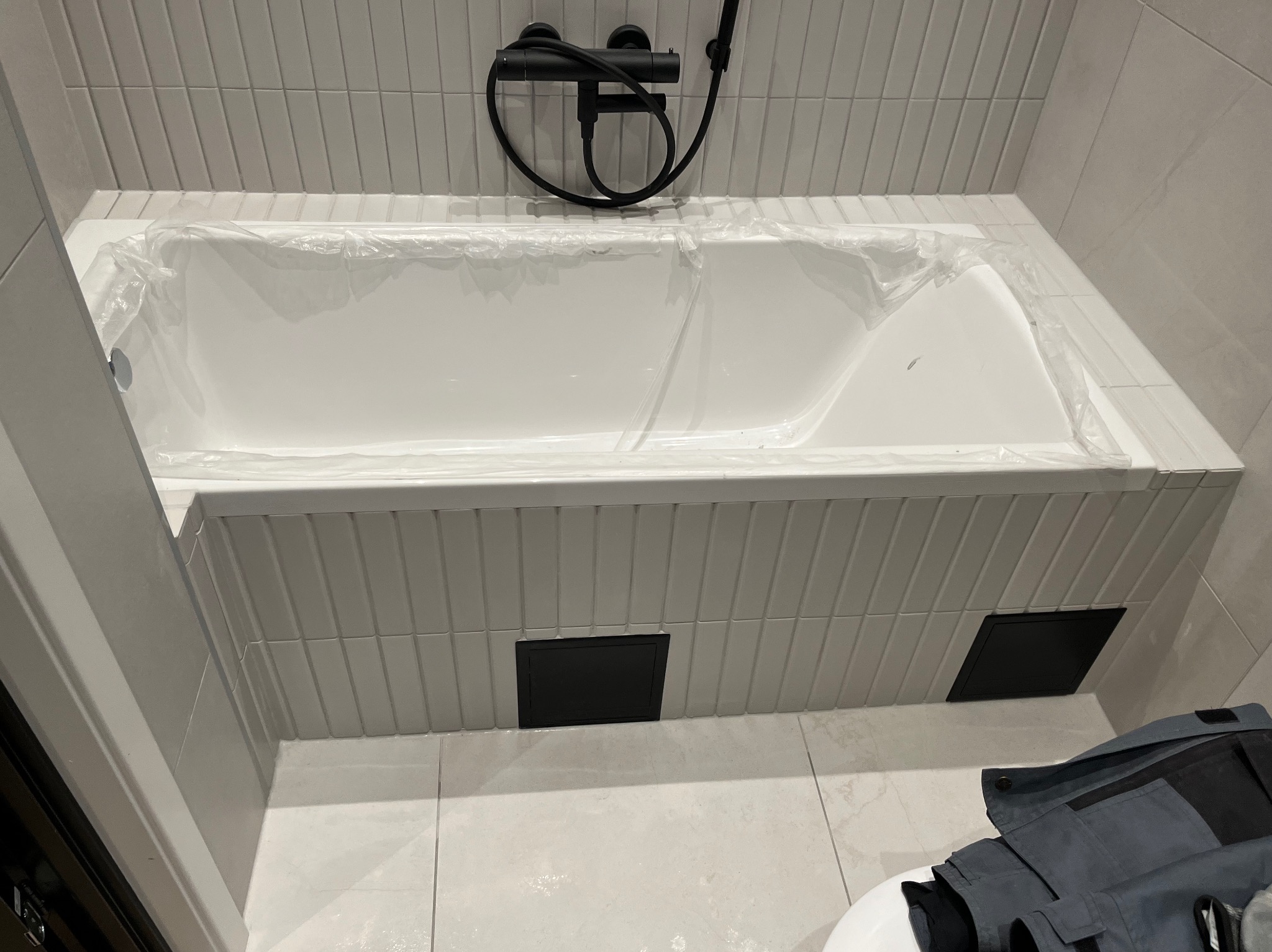 Biała wanna po montażu, zabezpieczona folią ochronną, z czarną armaturą. Obudowa wanny wykończona szarą, pionową glazurą. Dwa czarne otwory rewizyjne.