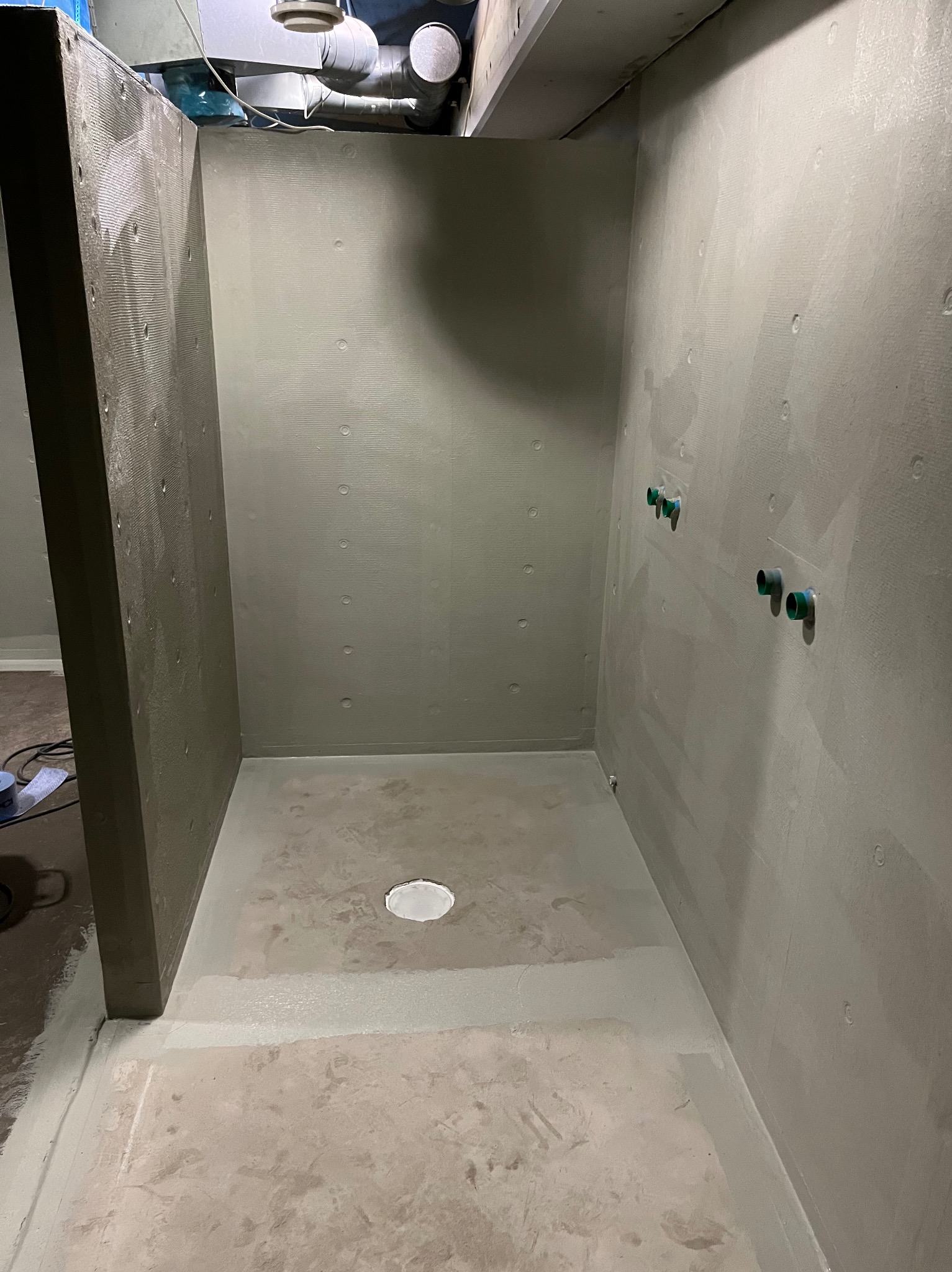 Przygotowana do wykończenia kabina prysznicowa z hydroizolacją ścian i podłogi, widoczne wyprowadzenia instalacji wodnej. Ujęcie z góry, szare tonacje.