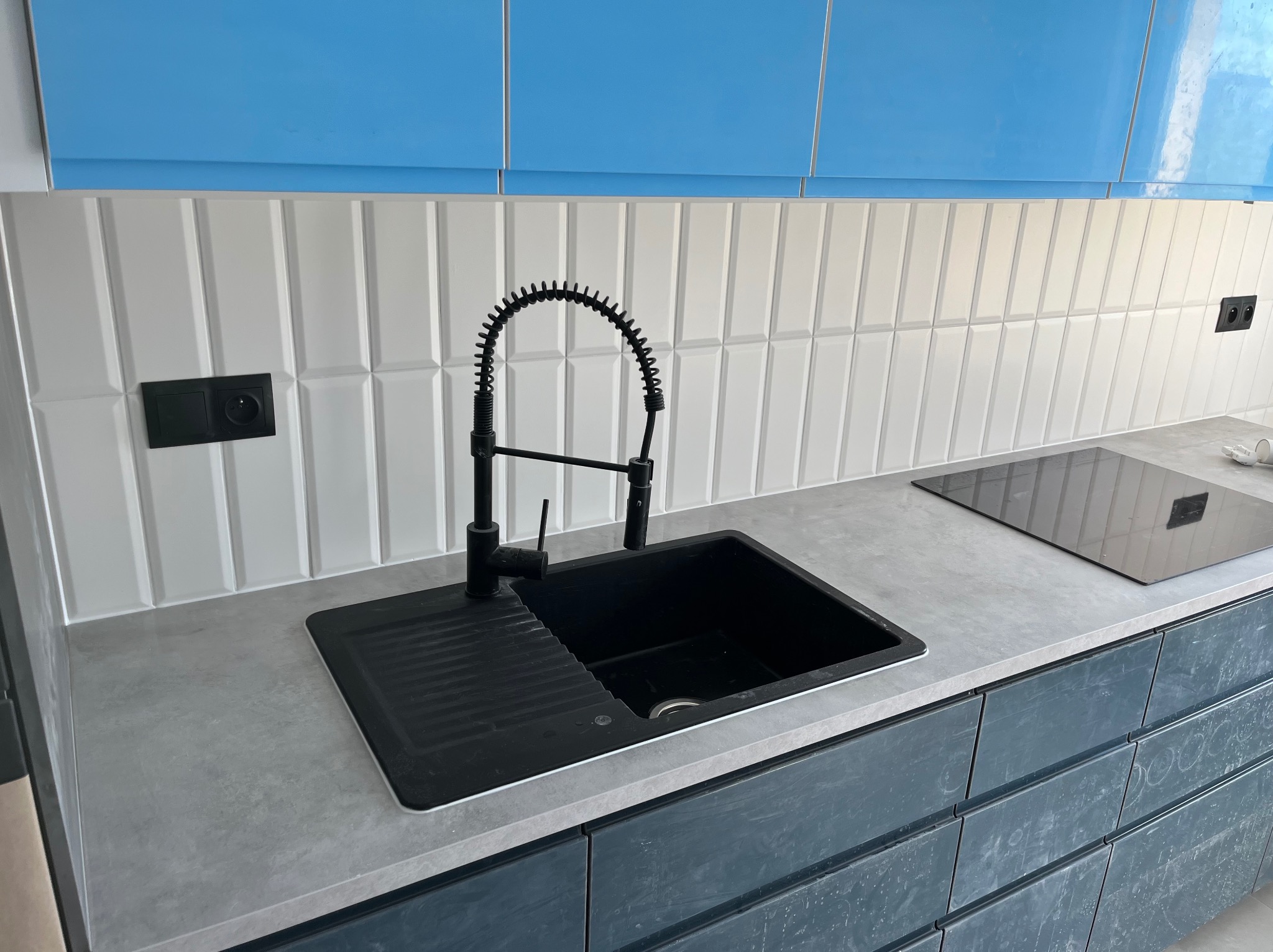 Nowoczesna kuchnia z czarnym zlewozmywakiem i baterią na tle białych, podłużnych płytek. Błękitne szafki górne.  Szare blaty i ciemne szafki dolne.