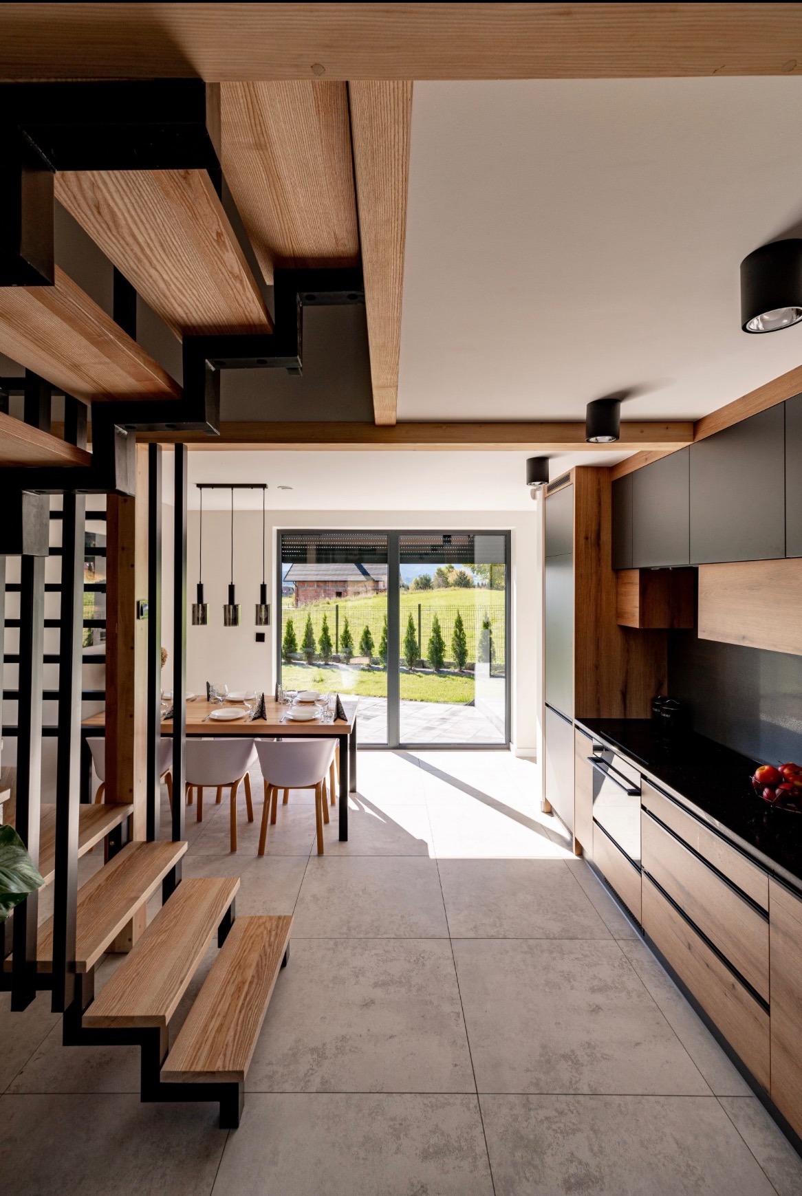 Nowoczesne wnętrze domu z drewnianymi schodami i kuchnią w odcieniach drewna i czerni. Widok na ogród przez duże okno, jasne płytki podłogowe.