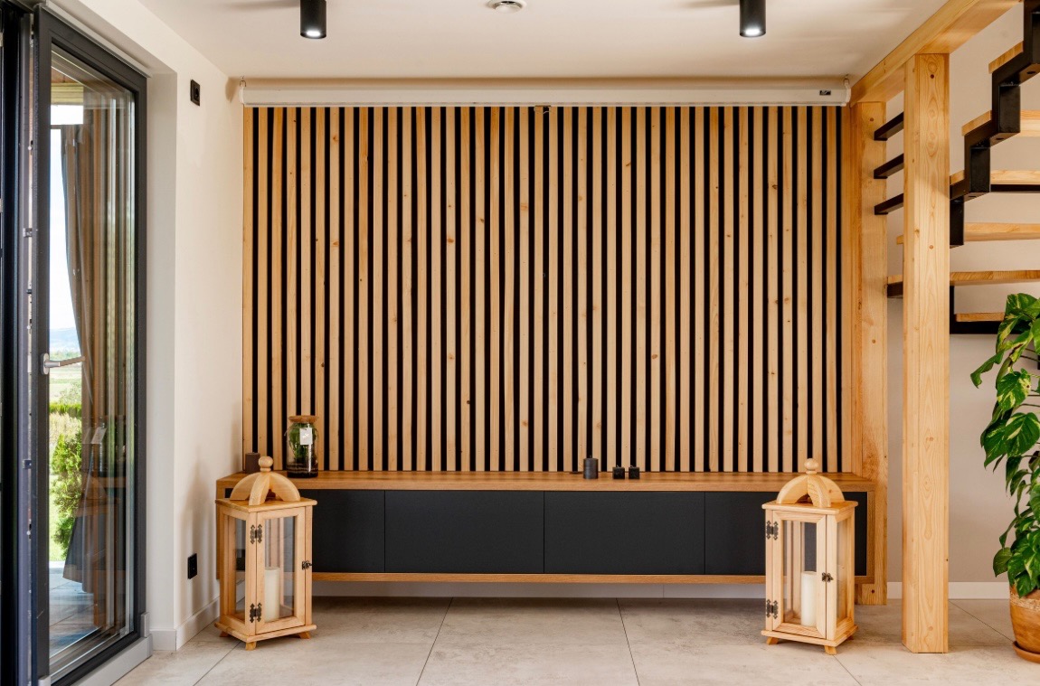 Nowoczesny salon z drewnianą ścianą z lameli, czarną komodą i drewnianymi lampionami. Widoczne schody z drewnianymi stopniami i zielona roślina doniczkowa.