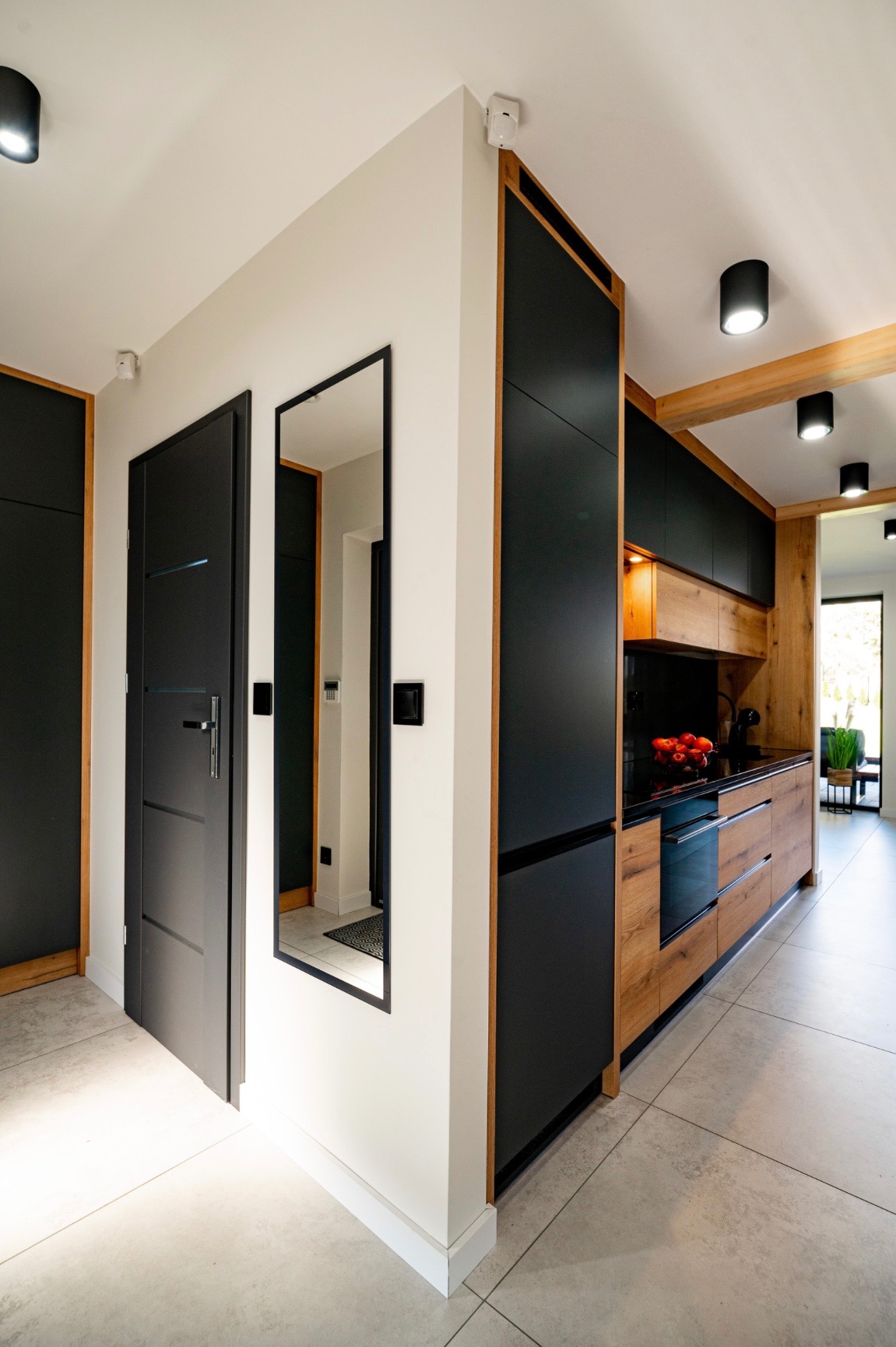 Nowoczesne wnętrze: czarne drzwi z lustrem, drewniana zabudowa kuchenna z czarnymi frontami, jasna podłoga i oświetlenie punktowe tworzą spójną całość.