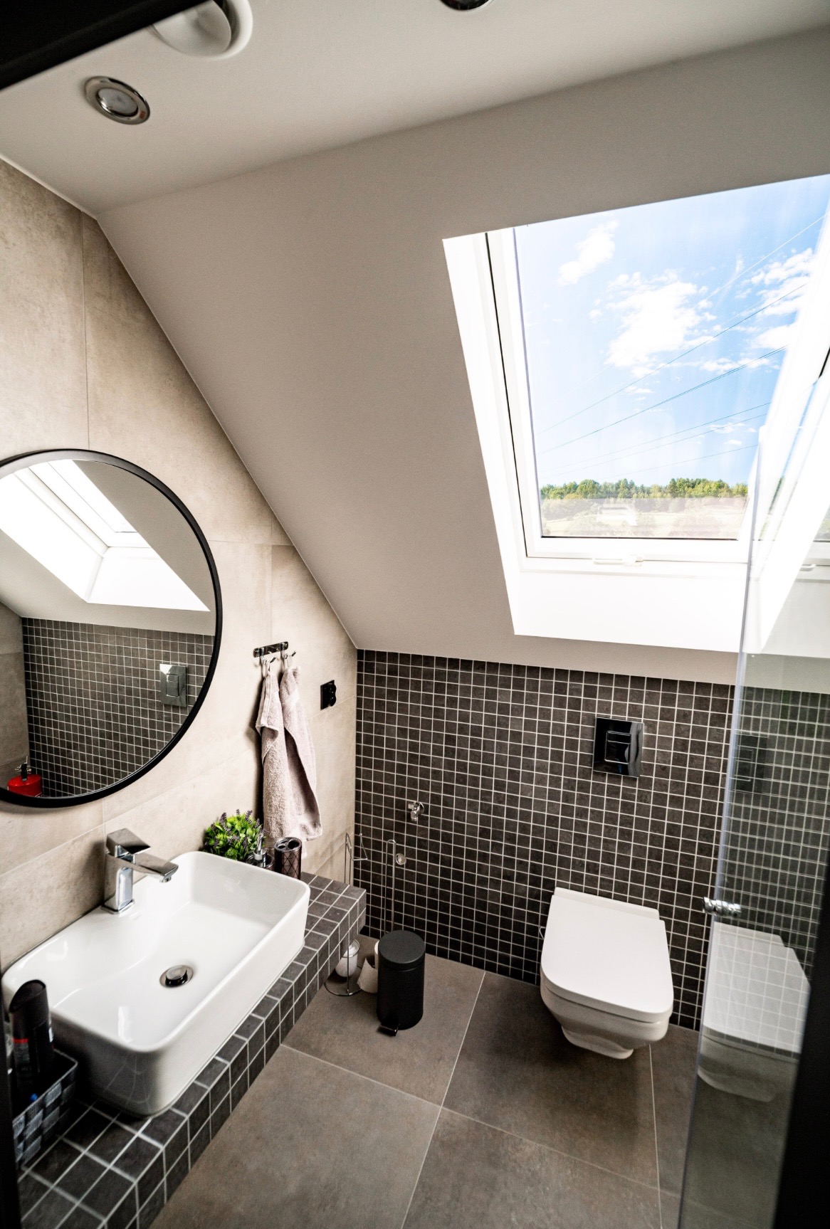 Stylowa łazienka na poddaszu z oknem dachowym i mozaiką. Okrągłe lustro odbija światło, a nowoczesna umywalka i toaleta dopełniają aranżację.