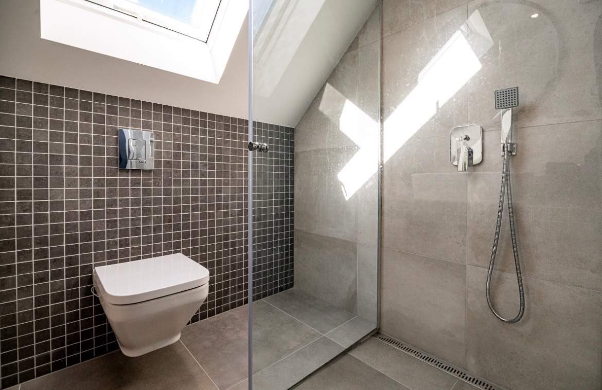 Nowoczesna łazienka z kabiną prysznicową typu walk-in, szarą mozaiką i podwieszaną toaletą. Okno dachowe wpuszcza naturalne światło.