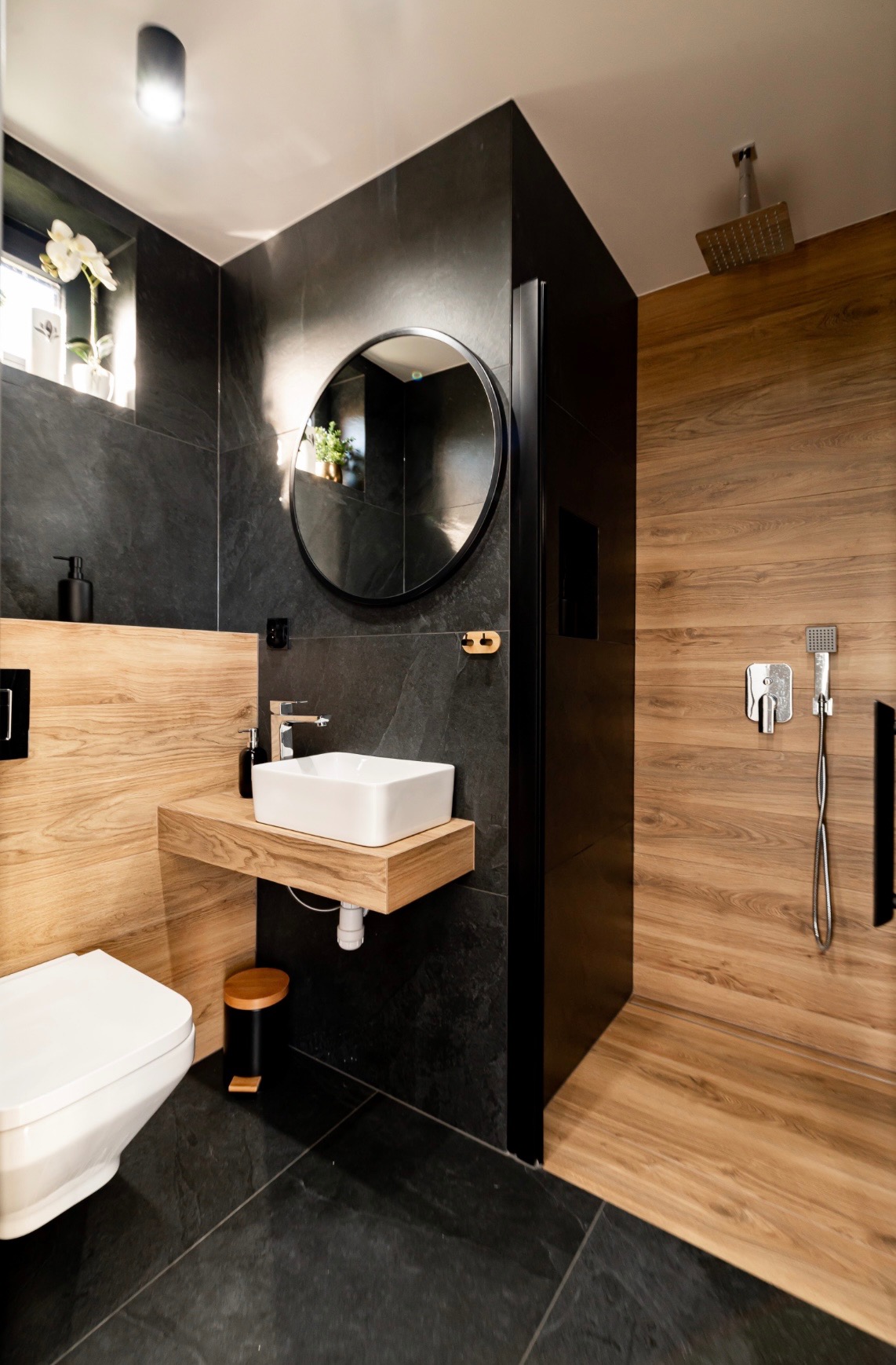 Nowoczesna łazienka z czarnymi płytkami, drewnopodobnymi panelami i okrągłym lustrem. Uwagę zwraca minimalistyczna umywalka na drewnianym blacie.