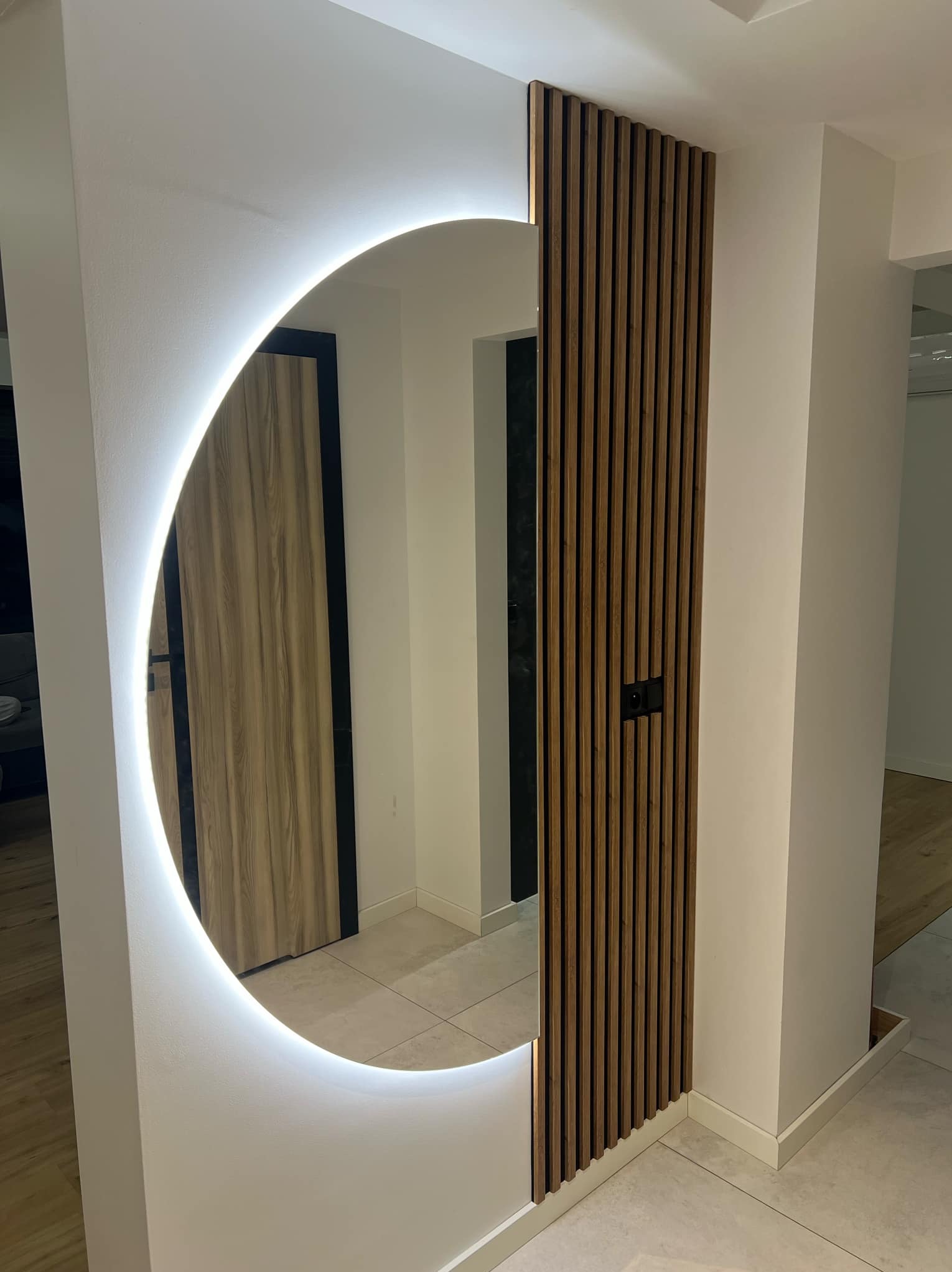 Nowoczesna aranżacja przedpokoju: okrągłe lustro z podświetleniem LED i drewniana ścianka z lameli, tworzące stylowy element dekoracyjny wnętrza.
