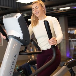 Anna Zawada Trener Personalny - Uśmiechnięta blondynka z aparatem na zębach ćwiczy na maszynie eliptycznej Matrix w siłowni. Widoczna butelka z wodą i ręcznik. Jasne oświetlenie.