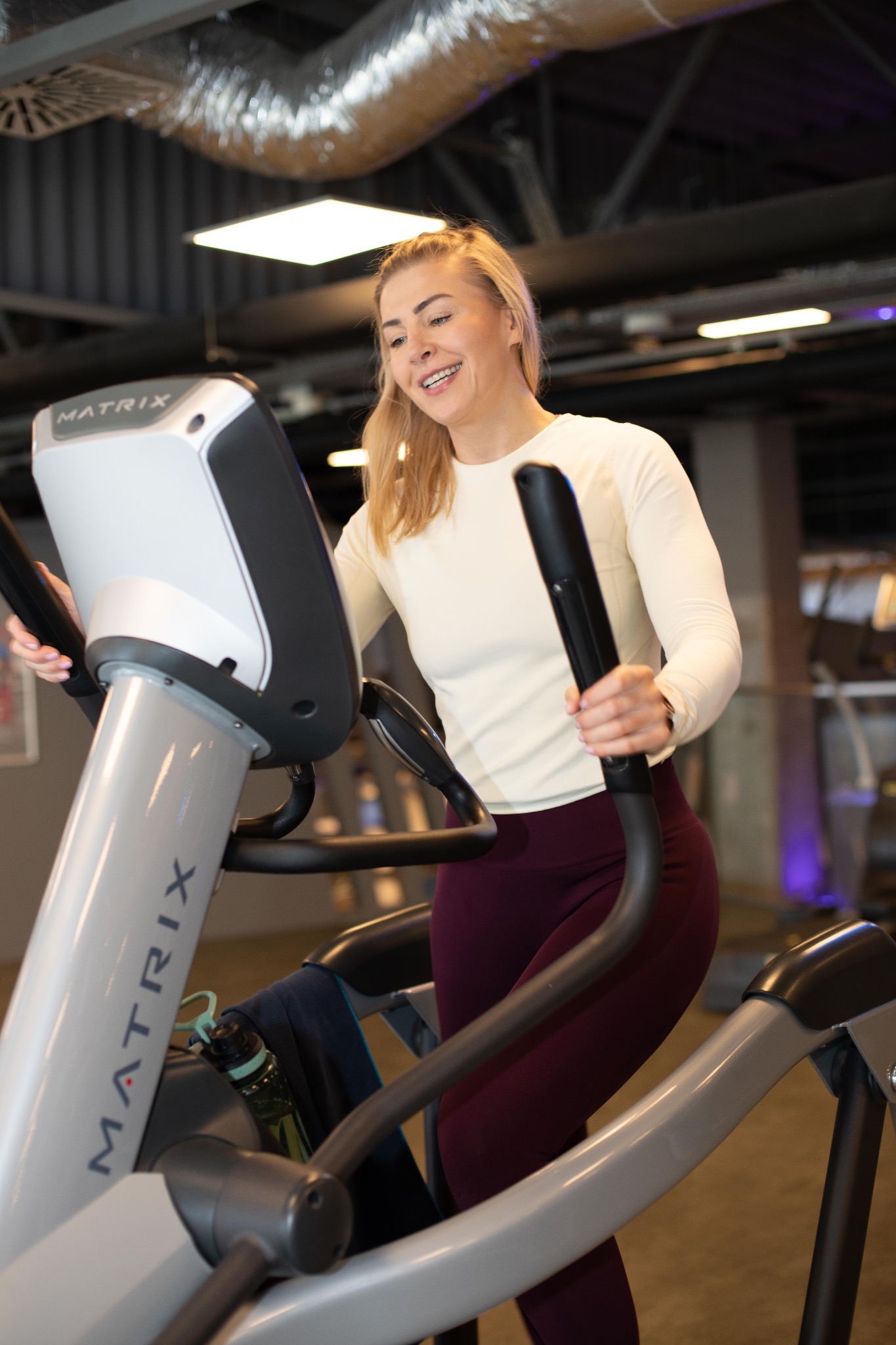 Uśmiechnięta blondynka z aparatem na zębach ćwiczy na maszynie eliptycznej Matrix w siłowni. Widoczna butelka z wodą i ręcznik. Jasne oświetlenie.