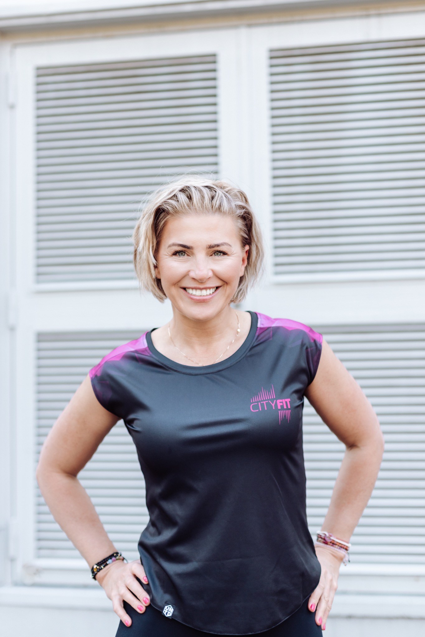 Uśmiechnięta blondynka w sportowej koszulce CITY FIT, z rękami na biodrach, pozuje na tle białych, zamkniętych okiennic.