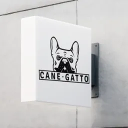Kanciasty szyld z wizerunkiem buldoga francuskiego i napisem 'CANE-GATTO' na szarej, betonowej ścianie. Detal minimalistycznego oznakowania firmy.