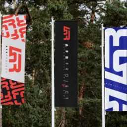 Trzy flagi reklamowe AFR Project z logo w kolorach czerwonym, czarnym i niebieskim, umieszczone na tle zielonych drzew.