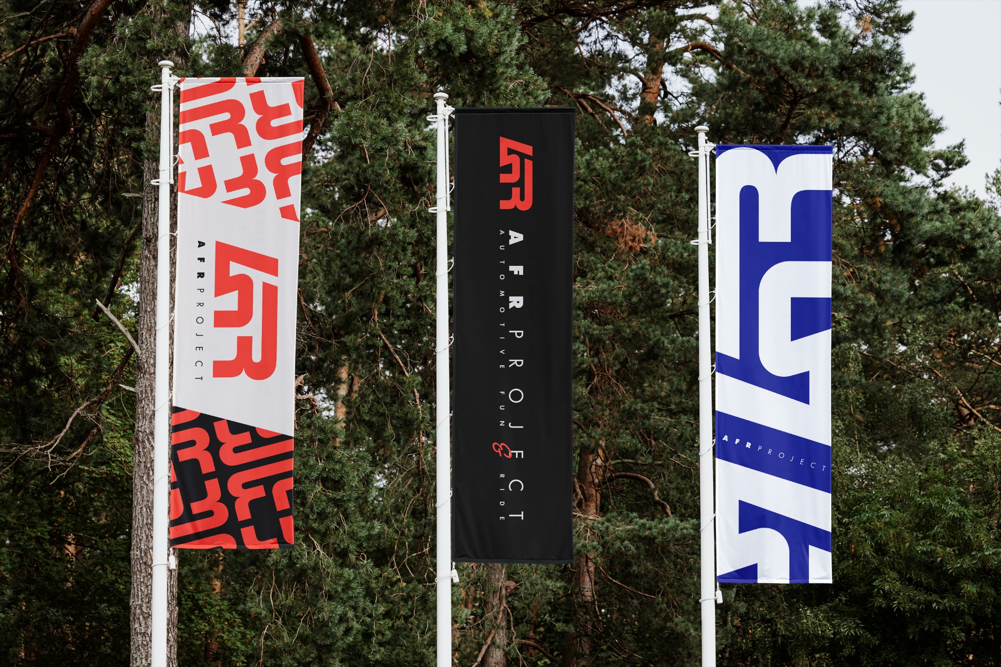 Trzy flagi reklamowe AFR Project z logo w kolorach czerwonym, czarnym i niebieskim, umieszczone na tle zielonych drzew.
