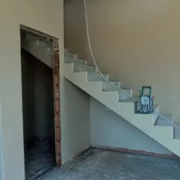 Wnętrze w trakcie wykańczania: ściany pokryte świeżym tynkiem, widoczne schody, otwór drzwiowy z ceglanym obramowaniem i narzędzie pomiarowe na schodach.