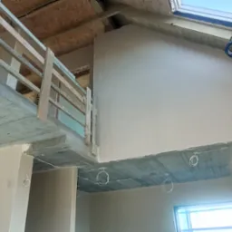 Wnętrze w trakcie wykańczania: gładkie ściany, betonowy strop z instalacją, drewniana balustrada. Widoczne okno dachowe i fragment okna. Jasne, naturalne światło.