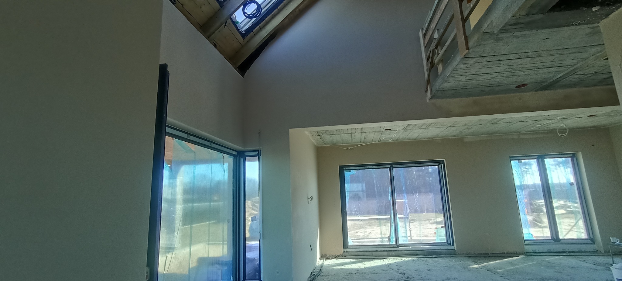 Wnętrze domu w trakcie prac wykończeniowych: gładkie ściany, duże okna z folią ochronną, surowy strop i drewniana konstrukcja antresoli. Widoczne instalacje.