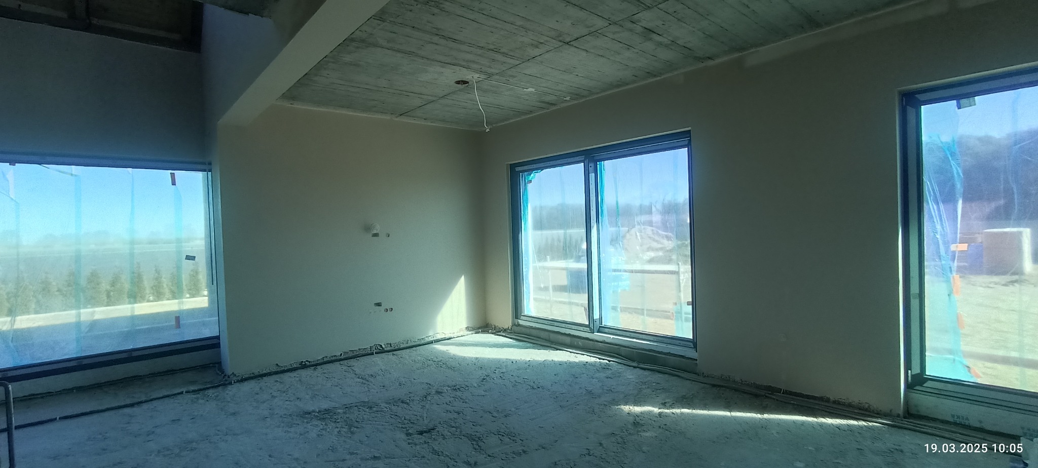 Surowe wnętrze z oknami zabezpieczonymi folią, ściany przygotowane do dalszych prac wykończeniowych, sufit betonowy. Widoczne instalacje na podłodze.