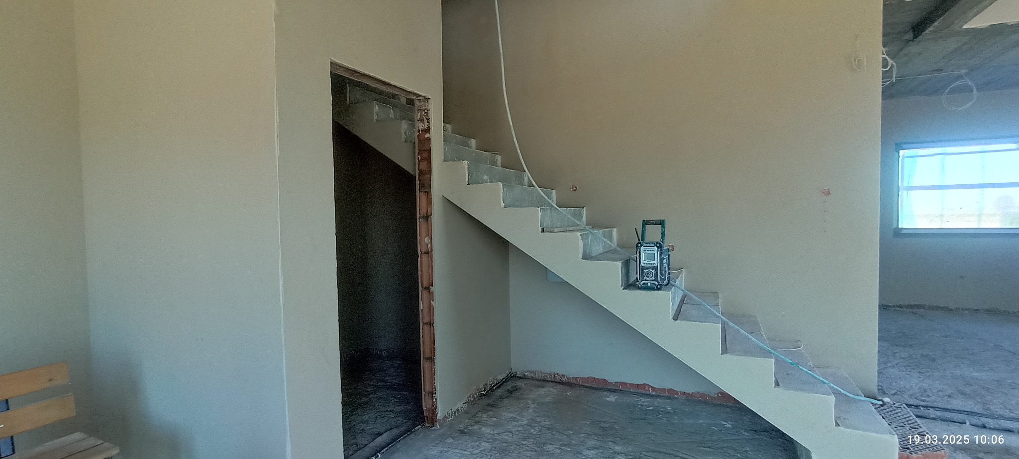 Wnętrze w trakcie wykańczania: ściany pokryte świeżym tynkiem, widoczne schody, otwór drzwiowy z ceglanym obramowaniem i narzędzie pomiarowe na schodach.