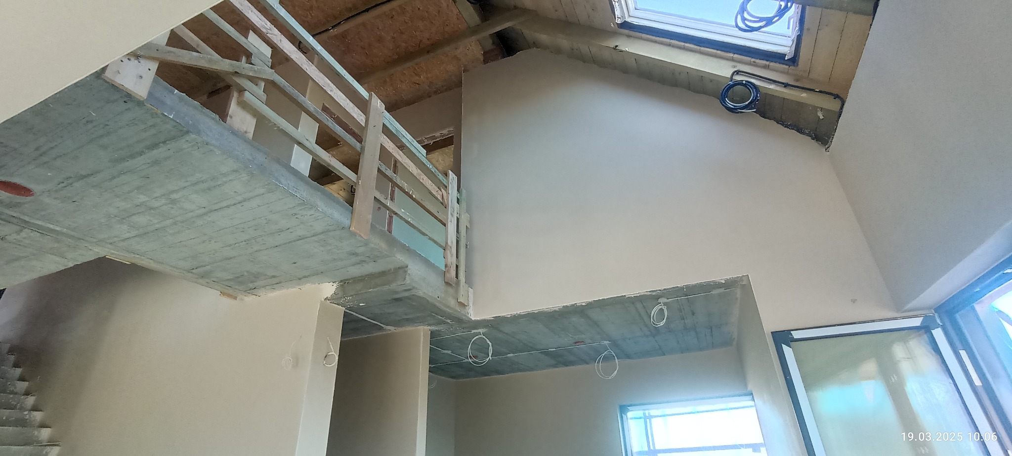Wnętrze w trakcie wykańczania: gładkie ściany, betonowy strop z instalacją, drewniana balustrada. Widoczne okno dachowe i fragment okna. Jasne, naturalne światło.