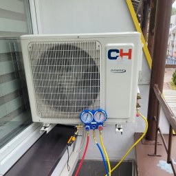 NEVCO Aleksander Kristof - Biała jednostka zewnętrzna klimatyzacji typu split, zamontowana na balkonie, podłączona do manometrów serwisowych z kolorowymi przewodami.