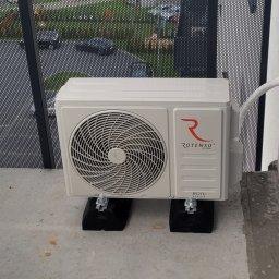 NEVCO Aleksander Kristof - Biała jednostka zewnętrzna klimatyzacji Rotenso Roni Series zamontowana na balkonie, z widocznym przewodem wchodzącym w ścianę. Tło stanowi ażurowa balustrada.