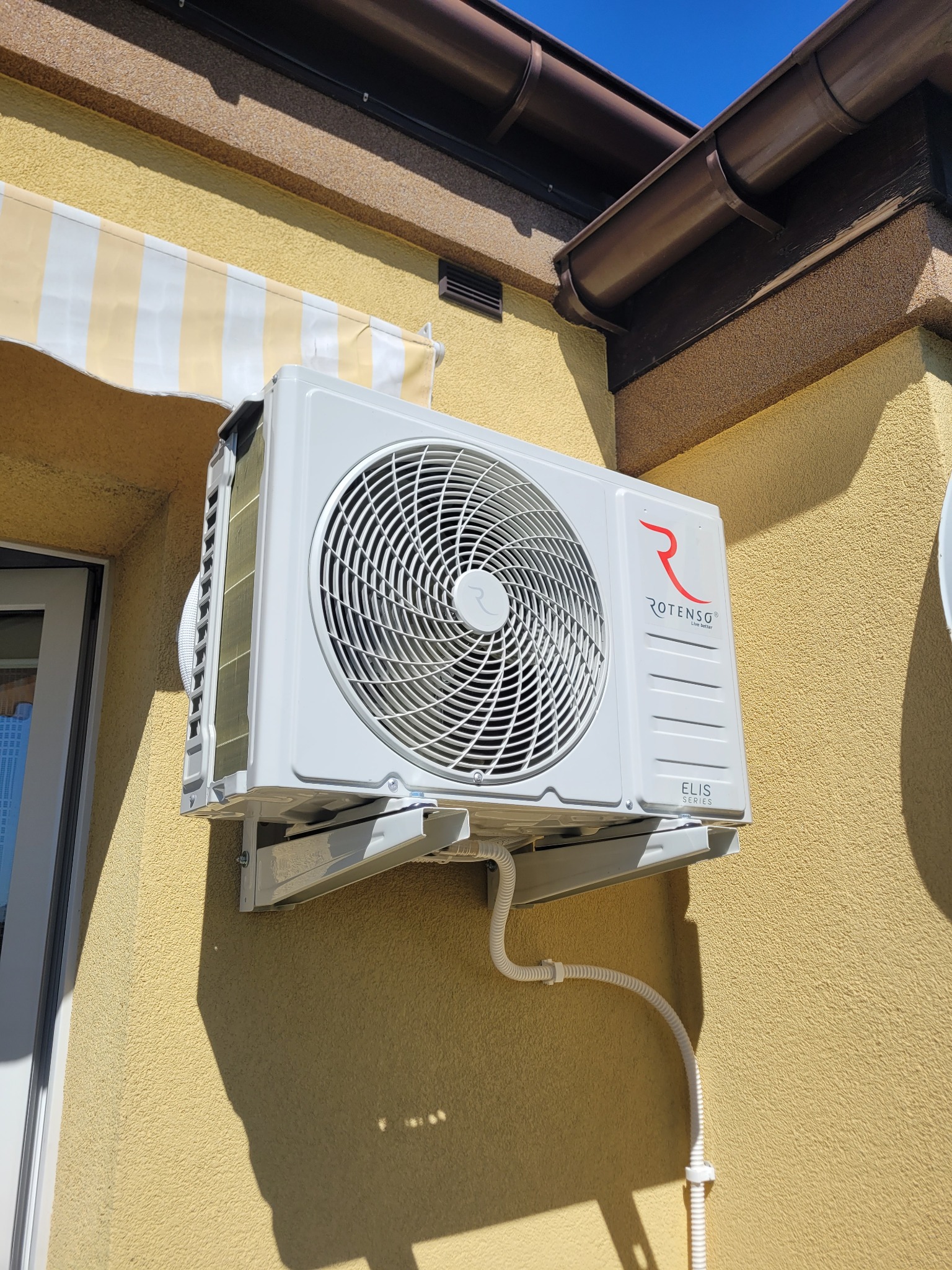 Biała klimatyzacja Rotenso Elis Series zamontowana na żółtej ścianie budynku z widocznym fragmentem okna i markizy w słoneczny dzień.