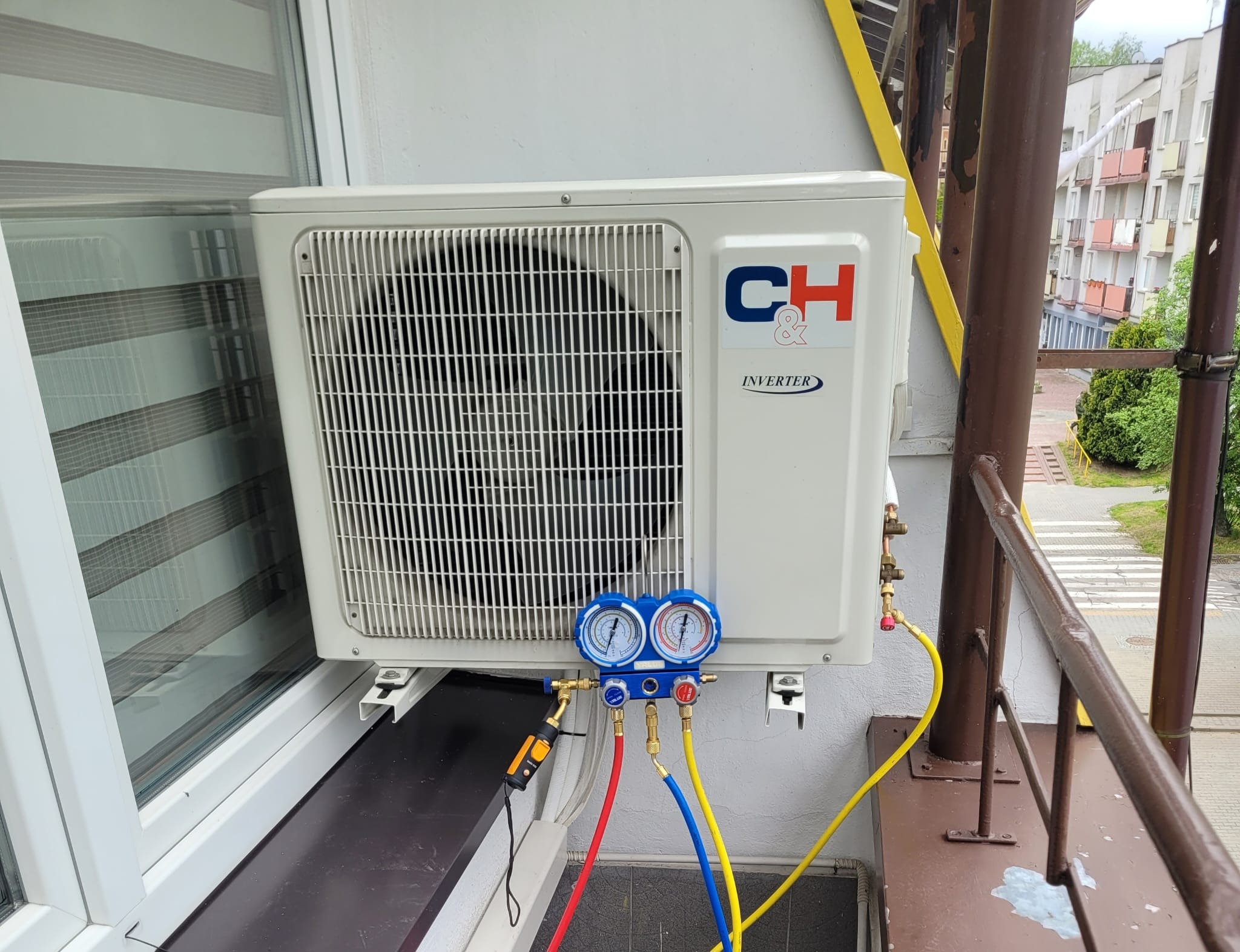 Biała jednostka zewnętrzna klimatyzacji typu split, zamontowana na balkonie, podłączona do manometrów serwisowych z kolorowymi przewodami.