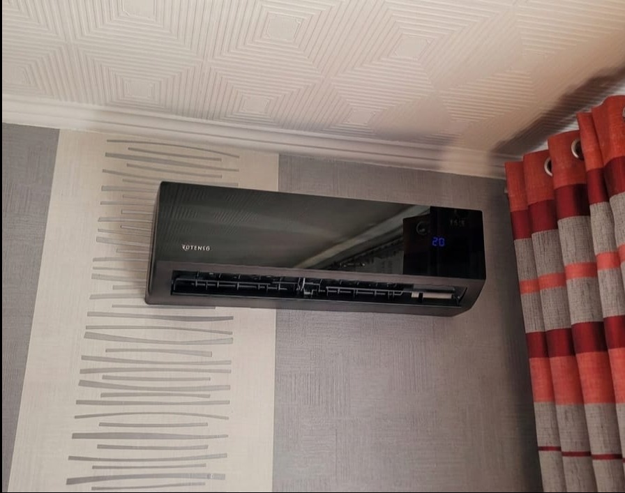 Czarny klimatyzator Rotenso na ścianie z wzorzystą tapetą, obok zasłony w odcieniach czerwieni. Widoczny wyświetlacz z temperaturą 20 stopni.