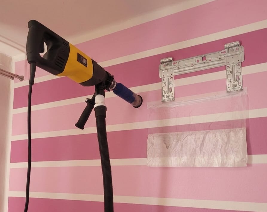 Wiercenie otworu w różowej ścianie w paski pod montaż klimatyzacji. Uchwyt montażowy na ścianie, worek na pył. Wiertarka z odsysaniem pyłu.