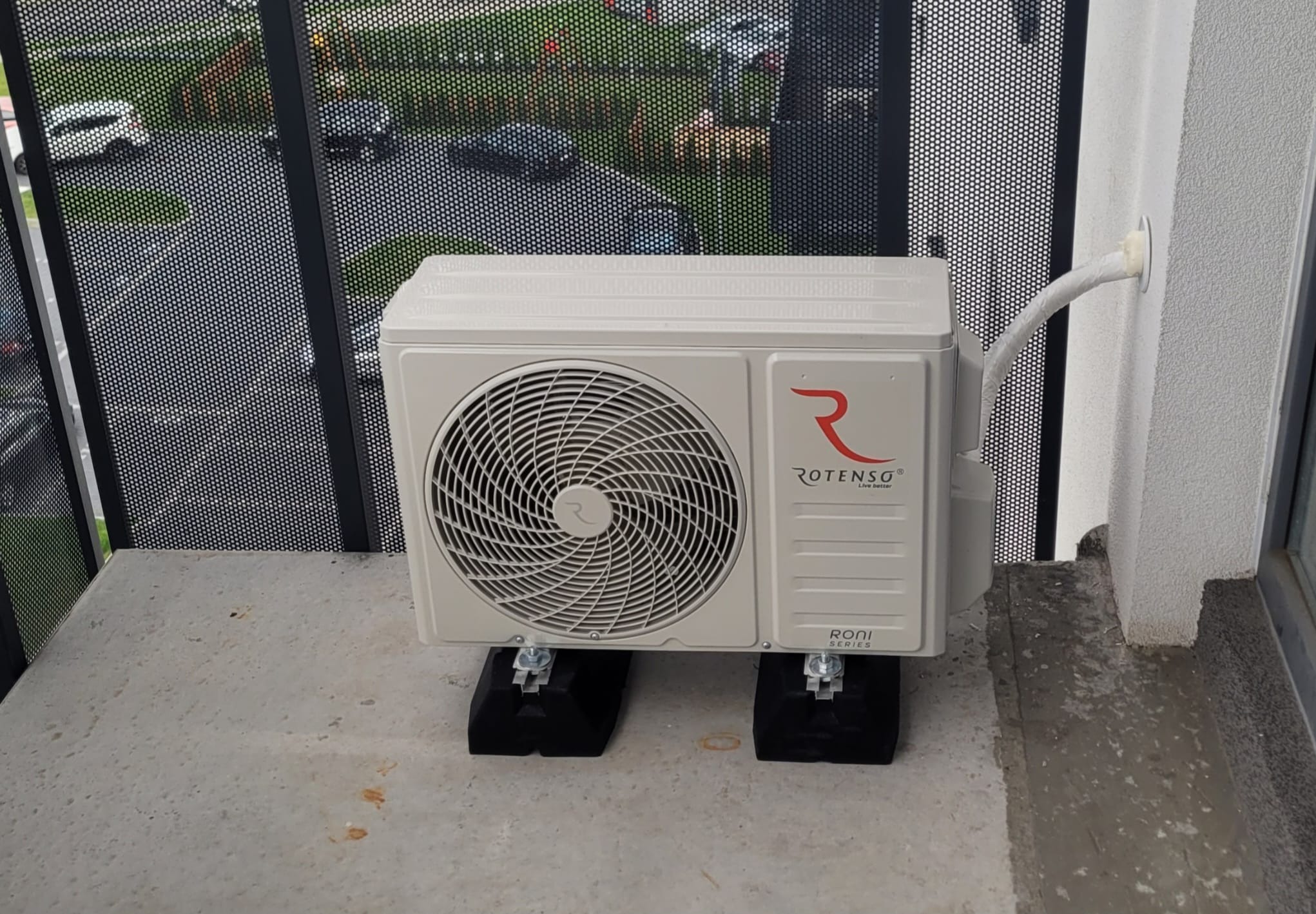 Biała jednostka zewnętrzna klimatyzacji Rotenso Roni Series zamontowana na balkonie, z widocznym przewodem wchodzącym w ścianę. Tło stanowi ażurowa balustrada.