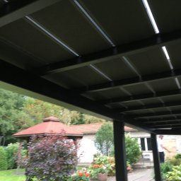 Expeert Polska sp. z o. o. - Zadaszenie tarasu z regulowanymi panelami, widok z dołu ukazujący konstrukcję i oświetlenie, w tle ogród z altaną i domem jednorodzinnym.