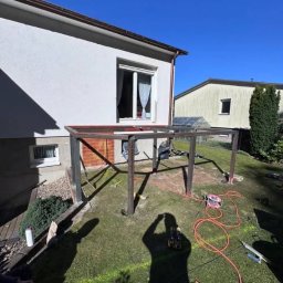 Expeert Polska sp. z o. o. - Metalowa konstrukcja zadaszenia tarasu w trakcie montażu przy domu jednorodzinnym, widoczne narzędzia i cień osoby robiącej zdjęcie. Prace montażowe w toku.