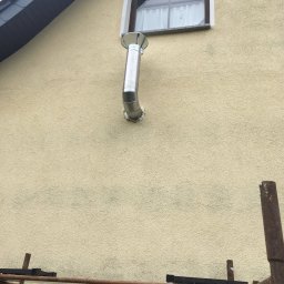 Expeert Polska sp. z o. o. - Elewacja budynku z zamontowanym metalowym kominem, widoczne rusztowanie. Kremowa ściana z oknem i fragment dachu. Detal instalacji kominowej.