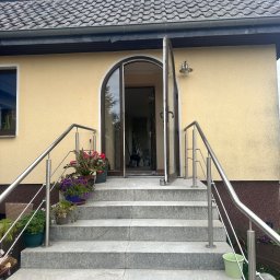Expeert Polska sp. z o. o. - Wejście do domu z granitowymi schodami, stalową balustradą i otwartymi drzwiami łukowymi. Kwiaty w donicach dodają koloru. Elewacja budynku w ciepłym odcieniu żółci.