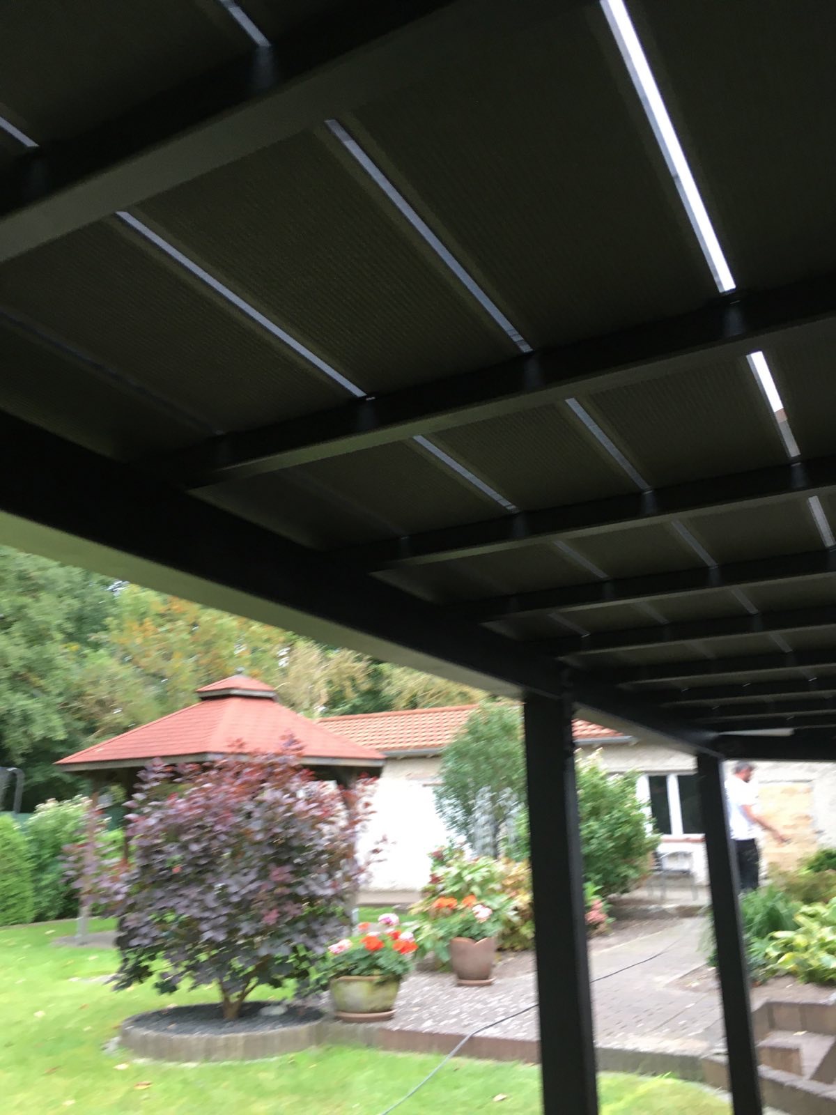 Zadaszenie tarasu z regulowanymi panelami, widok z dołu ukazujący konstrukcję i oświetlenie, w tle ogród z altaną i domem jednorodzinnym.