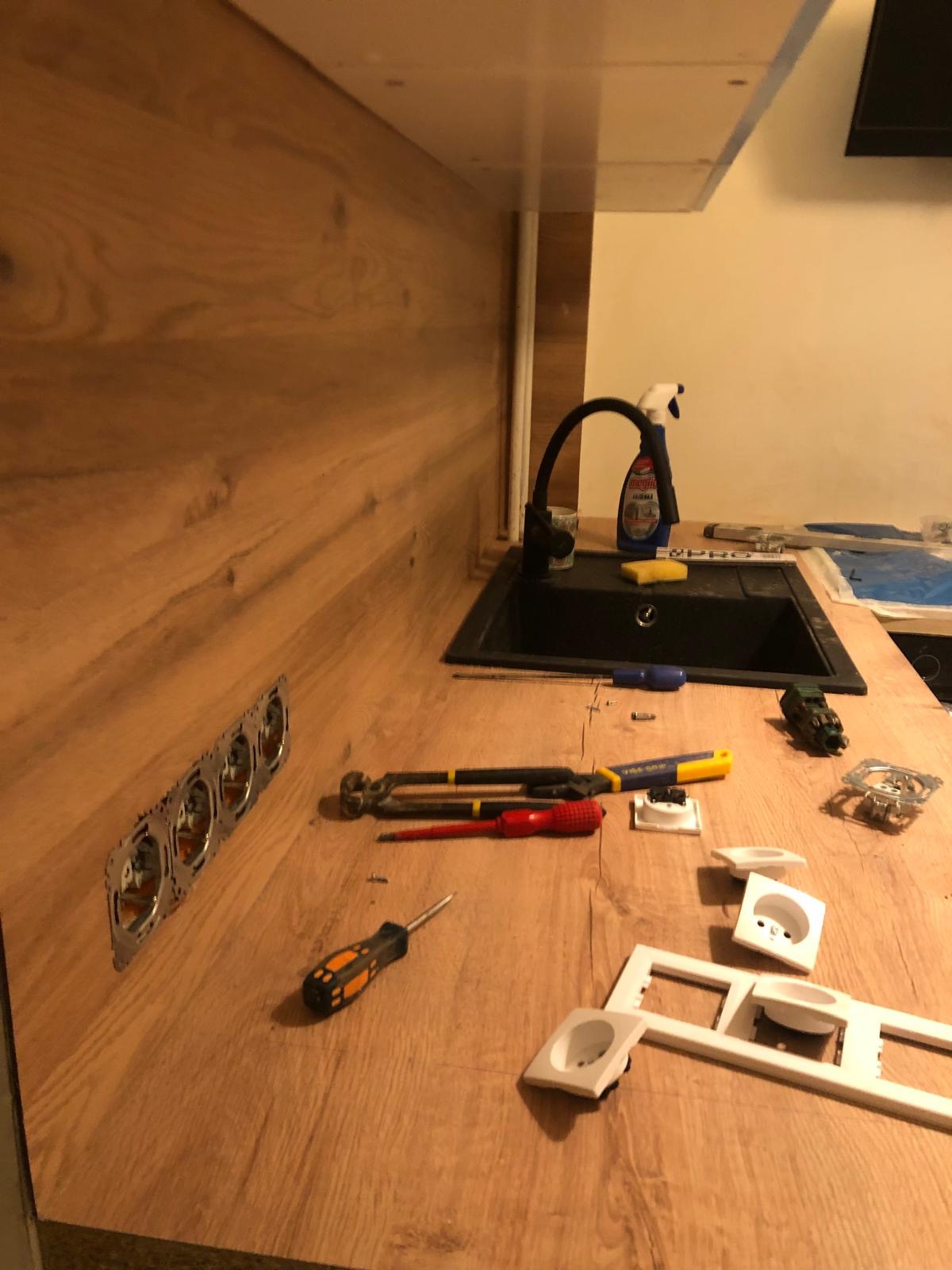 Montaż gniazdek elektrycznych w blacie kuchennym o drewnianym wykończeniu. Widoczne narzędzia, w tle zlew i armatura. Etap prac wykończeniowych.