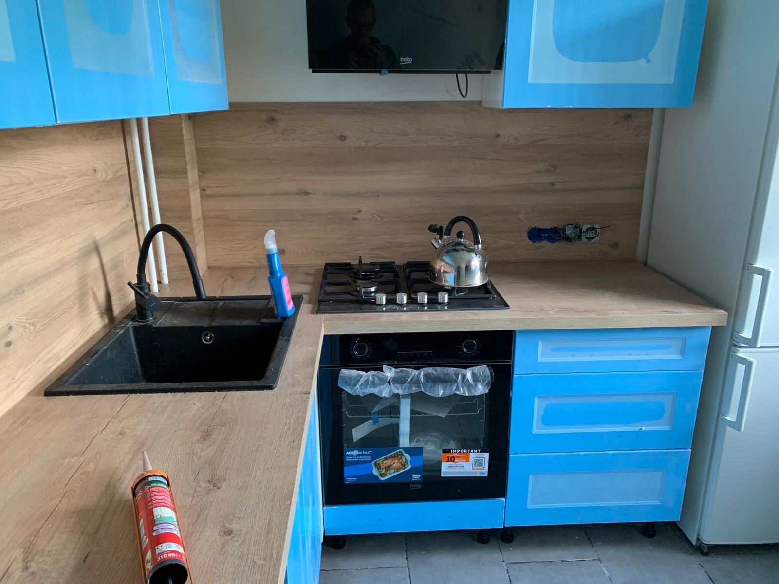 Jasna kuchnia z niebieskimi szafkami i drewnianym blatem. Widoczny zlew, kuchenka, piekarnik i akcesoria. W tle telewizor. Częściowo zabezpieczone fronty szafek.