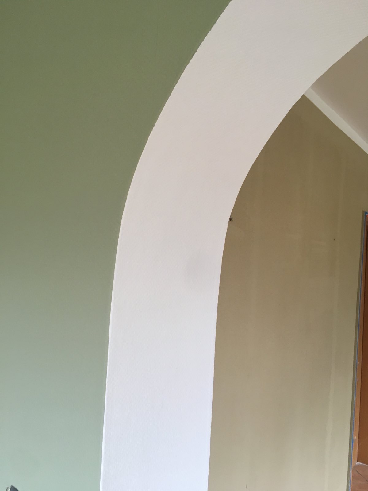 Oliwkowa ściana sąsiaduje z łukiem w kolorze złamanej bieli, tworząc kontrastowe połączenie barw. Fragment drzwi w tle po prawej stronie.