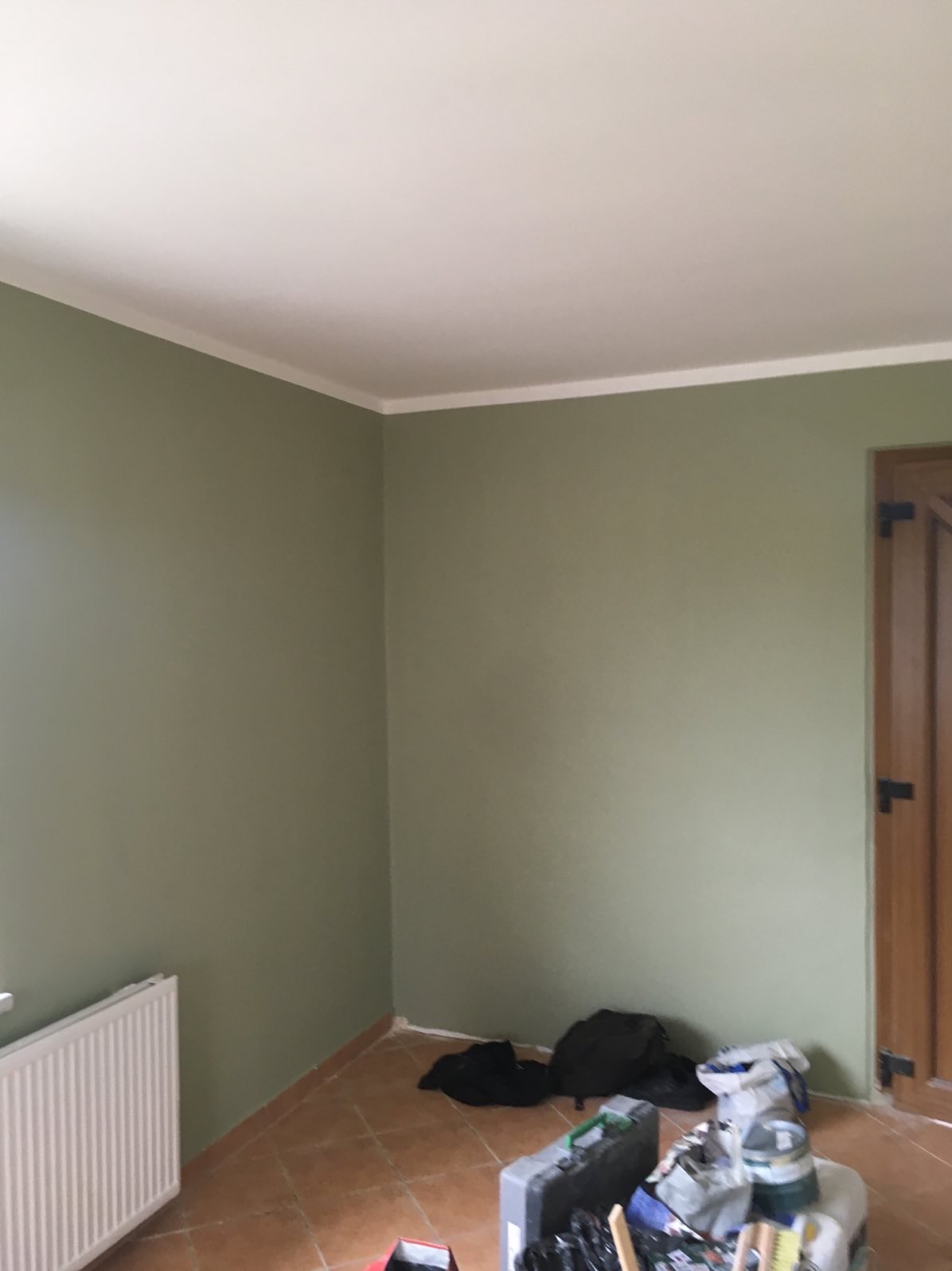 Pomieszczenie po malowaniu ścian na oliwkowy kolor, widoczne narzędzia malarskie, brązowa podłoga i biały sufit z listwą.