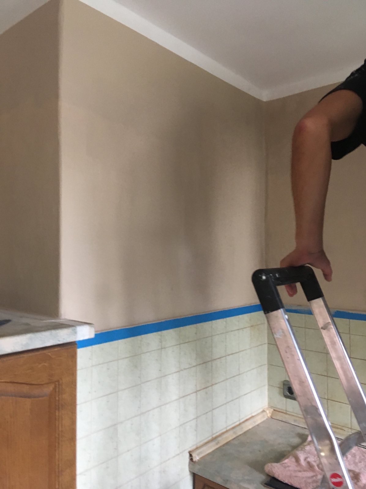 Ściana po malowaniu na beżowo w pomieszczeniu z kafelkami, zabezpieczonymi niebieską taśmą malarską. Fragment drabiny i ręka w kadrze.