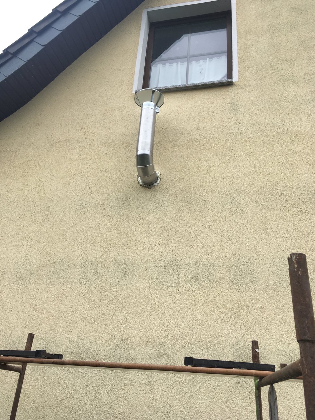 Elewacja budynku z zamontowanym metalowym kominem, widoczne rusztowanie. Kremowa ściana z oknem i fragment dachu. Detal instalacji kominowej.