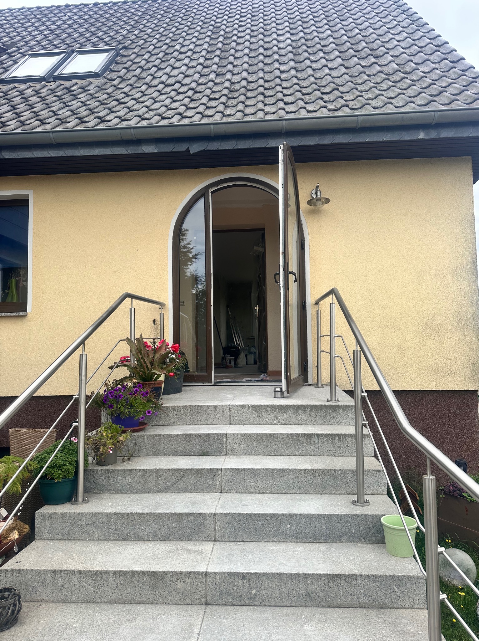 Wejście do domu z granitowymi schodami, stalową balustradą i otwartymi drzwiami łukowymi. Kwiaty w donicach dodają koloru. Elewacja budynku w ciepłym odcieniu żółci.