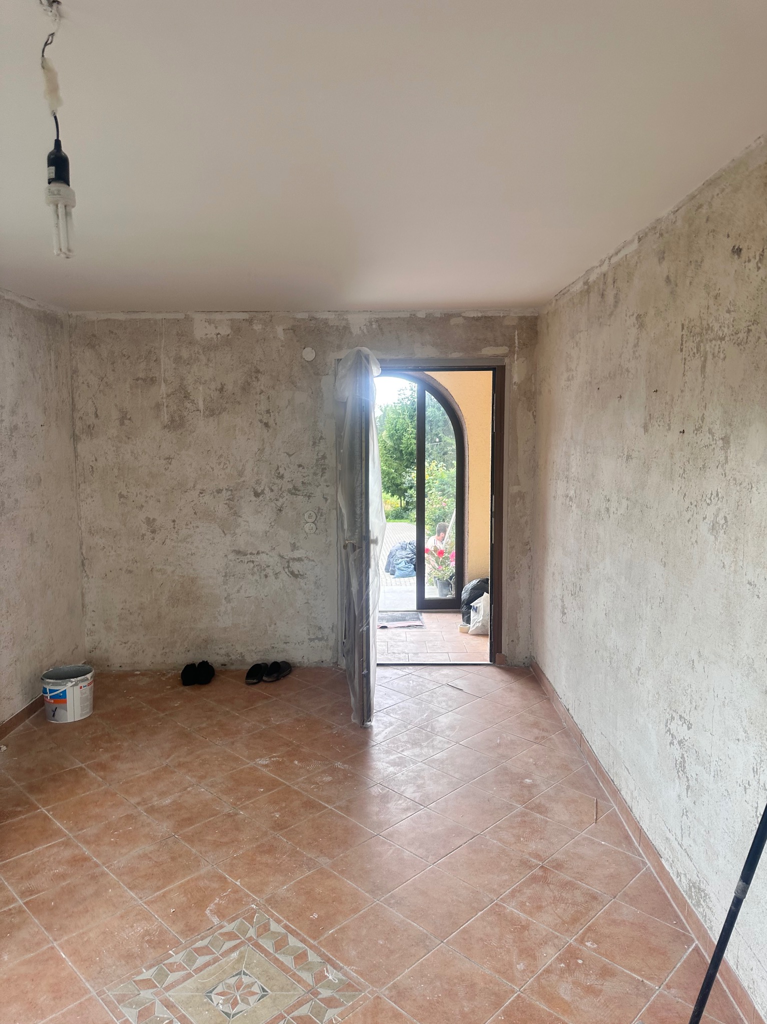 Pokój w trakcie prac wykończeniowych: surowe ściany, terakota na podłodze, widoczne ślady po remoncie. W tle otwarte drzwi na zewnątrz.