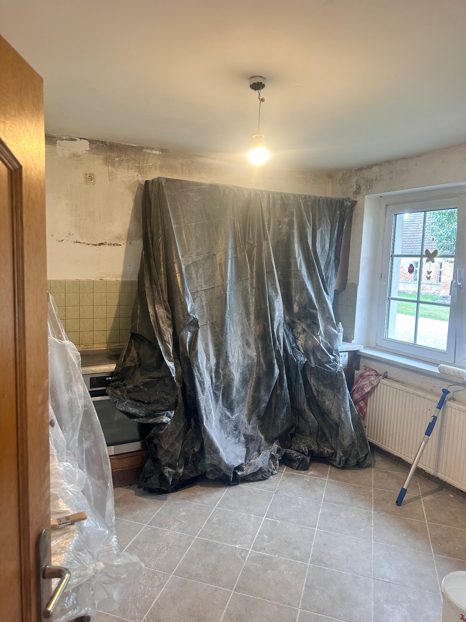 Kuchenne wnętrze w trakcie prac remontowych: ściany przygotowane do malowania, meble zabezpieczone folią, widoczny wałek malarski i wiadro z farbą.