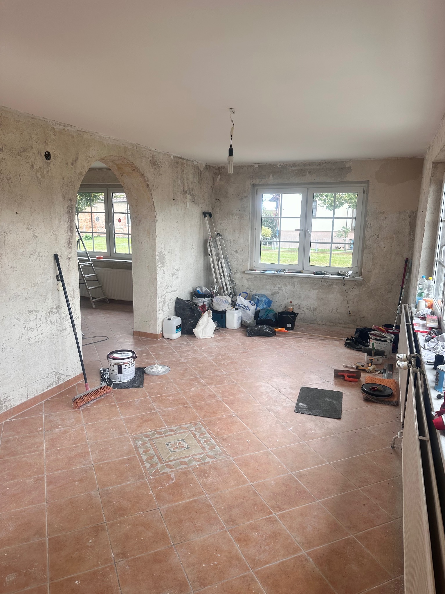 Pokój w trakcie prac remontowych: ściany przygotowane do malowania, widoczne narzędzia, drabina, puszki z farbą, podłoga wyłożona terakotą. Stan surowy.